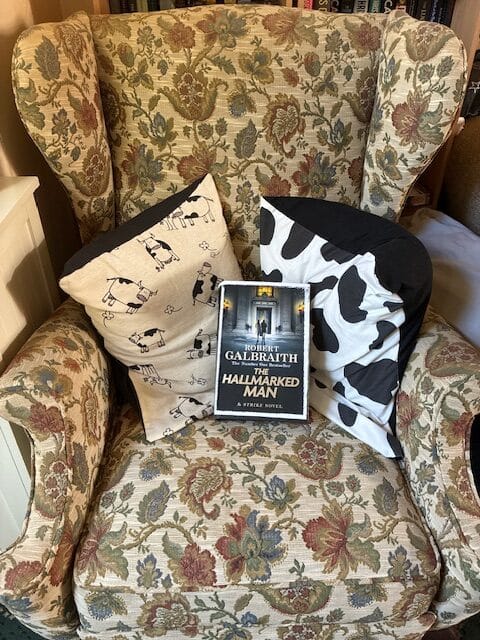 Cosy floral armchair with sheep and cow print cushions, featuring the crime novel The Hallmarked Man by Robert Galbraith placed in the centre, ready for an evening of thriller reading.