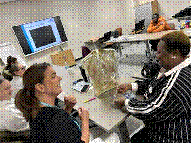Care staff working hands on with a clear PEG training torso during a Prima Cura Training practical session, with the feed preparation video on the screen behind them.